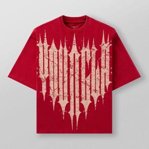 Puede incluir: Camiseta roja de manga corta con un gráfico grande, desgastado y blanquecino. El gráfico presenta un diseño de texto vertical estilizado con bordes afilados y puntiagudos. La camiseta tiene cuello redondo y corte holgado.