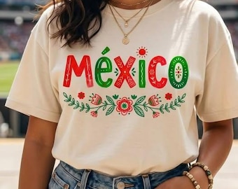 以下が含まれることがあります： 赤と緑の文字で「México」とプリントされたクリーム色の半袖Tシャツ。テキストの下には、赤と緑の装飾的な花や葉の模様が描かれています。Tシャツは青いデニムジーンズと組み合わされています。