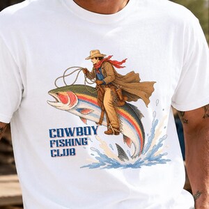 May include: A white short-sleeved t-shirt featuring a graphic illustration of a cowboy riding a large rainbow trout like a horse. The design includes the text "COWBOY FISHING CLUB" in blue lettering below the fish, with water splashing at the base.