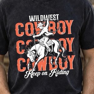 May include: Black t-shirt with a Wild West cowboy graphic. The design features a cowboy riding a horse, with the words "Wildest Cowboy" in red and white. The phrase "Keep on Riding" is also included.