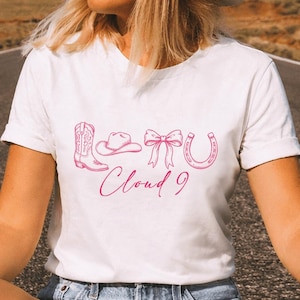 May include: A white short-sleeved t-shirt featuring a pink graphic print of a cowboy boot, a cowboy hat, a bow, and a horseshoe, with the text Cloud 9 written in a cursive script font below the icons, centered on the chest of the garment.