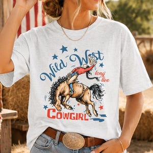 Puede incluir: Una camiseta gris claro de manga corta con un gráfico de una vaquera montando un caballo que corcovea. El texto dice Wild West y long live COWGIRL en azul y rojo, con estrellas azules. La camiseta se combina con pantalones cortos de mezclilla.