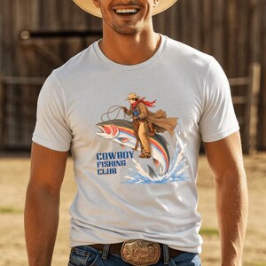 May include: A white short-sleeved t-shirt featuring a graphic of a cowboy riding a large rainbow trout like a horse. The illustration includes blue water splashes and the text COWBOY FISHING CLUB printed in bold blue letters below the fish.