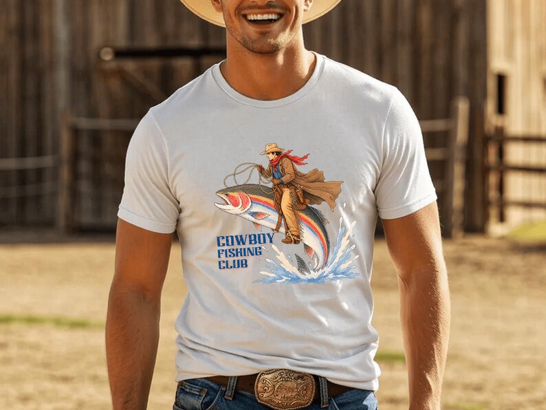 May include: A white short-sleeved t-shirt featuring a graphic of a cowboy riding a large rainbow trout like a horse. The illustration includes blue water splashes and the text COWBOY FISHING CLUB printed in bold blue letters below the fish.