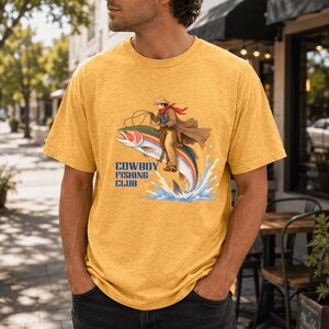 May include: A heathered mustard yellow short-sleeve t-shirt featuring a graphic illustration of a cowboy riding a large rainbow trout. The text "COWBOY FISHING CLUB" is printed in blue letters below the illustration, which shows water splashing.