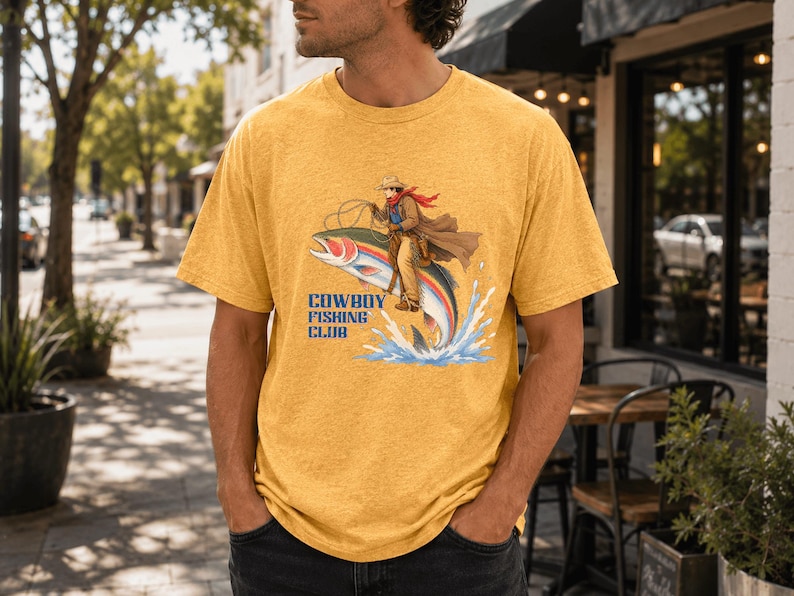 May include: A heathered mustard yellow short-sleeve t-shirt featuring a graphic illustration of a cowboy riding a large rainbow trout. The text "COWBOY FISHING CLUB" is printed in blue letters below the illustration, which shows water splashing.