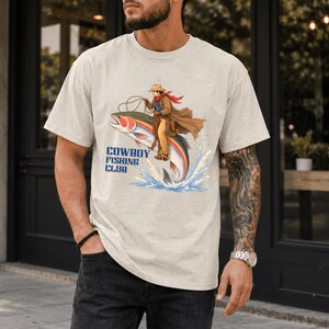 May include: A beige short-sleeve t-shirt featuring a graphic print of a cowboy riding a large rainbow trout like a horse. The cowboy wears a hat, brown coat, and red scarf. Below the illustration, the text reads COWBOY FISHING CLUB in blue.