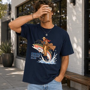 May include: A navy blue short-sleeve t-shirt featuring a graphic illustration of a cowboy riding a large rainbow trout. The cowboy wears a hat, duster, and red scarf. The text "COWBOY FISHING CLUB" is printed in blue letters below the fish.