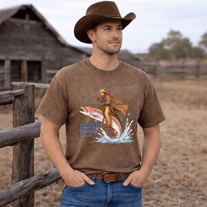 May include: A brown short-sleeve t-shirt featuring a graphic of a cowboy riding a large rainbow trout jumping out of water. The text "COWBOY FISHING CLUB" is printed in blue letters below the illustration. The shirt has a distressed, washed-out finish.