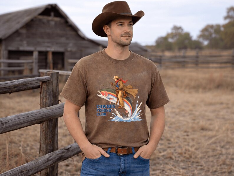May include: A brown short-sleeve t-shirt featuring a graphic of a cowboy riding a large rainbow trout jumping out of water. The text "COWBOY FISHING CLUB" is printed in blue letters below the illustration. The shirt has a distressed, washed-out finish.