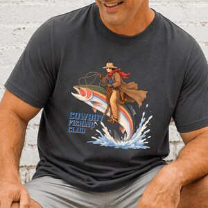 May include: A charcoal gray crew neck t-shirt featuring a graphic of a cowboy riding a large rainbow trout like a horse. The cowboy wears a hat, duster, and red scarf, holding a lasso. The text Cowboy Fishing Club is printed in blue letters.