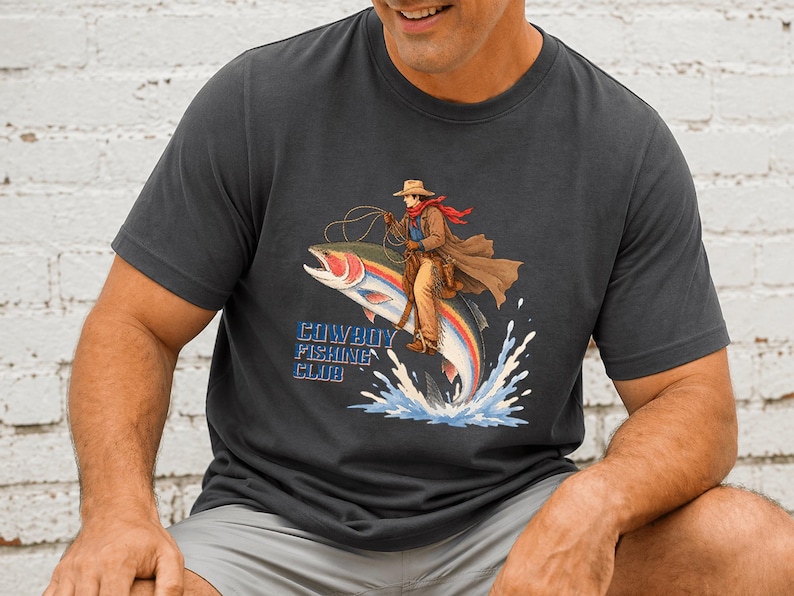 May include: A charcoal gray crew neck t-shirt featuring a graphic of a cowboy riding a large rainbow trout like a horse. The cowboy wears a hat, duster, and red scarf, holding a lasso. The text Cowboy Fishing Club is printed in blue letters.
