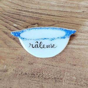 May include: A white, half-moon shaped ceramic dish with a blue rim and the word "râleuse" in black script. The dish is set against a wooden background.