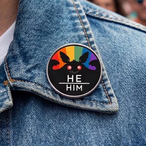 Könnte beinhalten: Ein schwarzer und regenbogenfarbener Anstecker mit dem Text "HE HIM" ist auf einer Jeansjacke befestigt. Der Anstecker zeigt ein Fledermaus-Design mit Regenbogenfarben auf schwarzem Hintergrund. Im Hintergrund sind eine Menschenmenge und Regenbogenfahnen zu sehen.