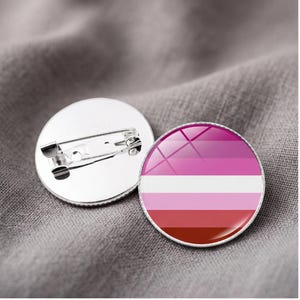 Puede incluir: Dos insignias redondas con el diseño de la bandera del orgullo lésbico. Las insignias presentan un degradado de rayas rosas, blancas y rojas. Una insignia muestra el diseño, mientras que la otra muestra la parte trasera.
