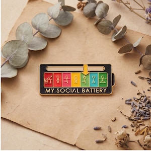 May include: A black and gold enamel pin with the text "MY SOCIAL BATTERY". The pin features a battery-shaped design with a series of colored bars and stick figures, indicating social energy levels. The arrow points to the green bar.
