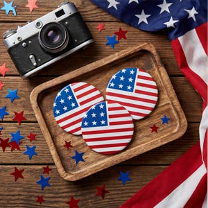 Könnte beinhalten: Eine Vintage-Kamera in Schwarz und Silber liegt in der Nähe eines Holztabletts mit drei runden Untersetzern mit dem Design der amerikanischen Flagge. Rote und blaue sternförmige Konfetti und ein Teil der amerikanischen Flagge vervollständigen die patriotische Szene.