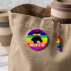 Könnte beinhalten: Ein runder Button mit Regenbogenhintergrund und einer schwarzen Katze mit Sonnenbrille. Der Text "ALLY CAT" steht unter der Katze. Ein Regenbogen-Perlen-Schlüsselanhänger hängt an einer beigefarbenen Tragetasche.