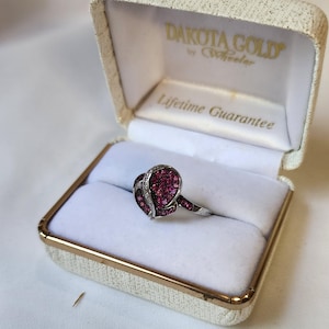 May include: A heart-shaped ring with a cluster of small, deep pink gemstones and a silver band, displayed in an open, cream-colored ring box with gold trim. The box is labeled "DAKOTA GOLD" and "Lifetime Guarantee".