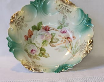 May include: A white porcelain bowl with a green and gold rim. The bowl is decorated with a floral design of pink and white roses.