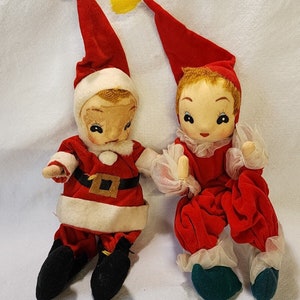 Puede incluir: Dos muñecas de duendes navideños vintage. Una está vestida de Papá Noel con un traje rojo, ribetes blancos y botas negras. El otro duende lleva un traje de terciopelo rojo con ribetes de encaje blanco y botas verdes.