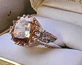 May include: A gold ring with a large, round, clear, faceted gemstone surrounded by smaller, clear, faceted gemstones. The ring is in a white ring box with a gold trim and the words "Lifetime" printed on the inside.