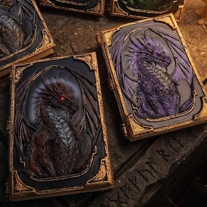Könnte beinhalten: Fünf rechteckige Bücher mit geprägten Drachen-Covern in Blau, Grün, Violett, Rot und Orange. Jedes Buch zeigt ein zentrales Drachenrelief, das von goldfarbenen Rändern umrahmt ist. Die Bücher sind auf einer Steinoberfläche neben drei Kerzen angeordnet.