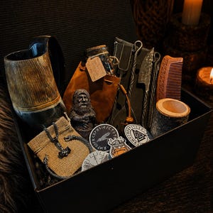 May include: A black gift box containing a horn mug, a brown leather pouch with a corked glass bottle, a small bearded figurine, a metal fork and knife set, a wooden comb, a cylindrical wood container, a metal pendant, and circular stickers.