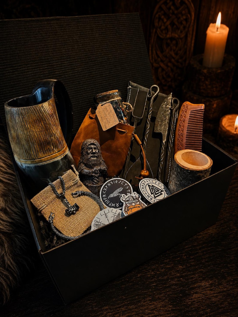 May include: A black gift box containing a horn mug, a brown leather pouch with a corked glass bottle, a small bearded figurine, a metal fork and knife set, a wooden comb, a cylindrical wood container, a metal pendant, and circular stickers.