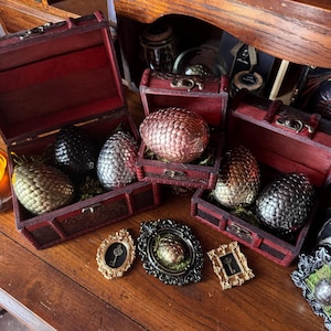 May include: Three dark red wooden chests containing textured, scale-patterned eggs in black, silver, copper, and gold finishes. Below the chests are three small, ornate frames, two containing key illustrations and one holding a small gold egg.