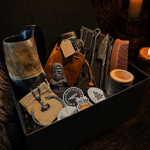 K&ouml;nnte beinhalten: Eine schwarze Geschenkbox mit einem Hornkrug, einem braunen Lederbeutel mit einer verkorkten Glasflasche, einer kleinen b&auml;rtigen Figur, einem Metallgabel- und Messerset, einem Holzkamm, einem zylindrischen Holzbeh&auml;lter, einem Metallanh&auml;nger und Aufklebern.
