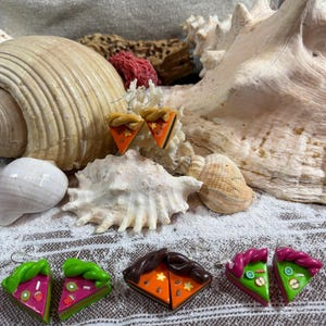 Può includere: Orecchini fatti a mano in pasta polimerica a forma di fette di torta. Gli orecchini presentano vari design, tra cui torta arancione con dettagli a stella e fette con frutta con accenti verdi, rosa e marroni. Esposti su una superficie strutturata con conchiglie.