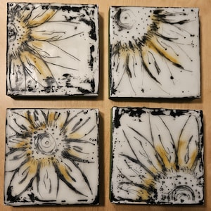 May include: Four square ceramic tiles with a sunflower design. Each tile features a white background with a black outline of a sunflower and yellow accents. The edges of the tiles are black, and the overall style is artistic and decorative.