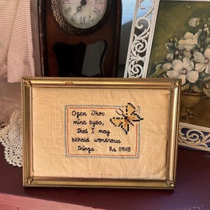 May include: A rectangular cross-stitch embroidery in a gold-toned metal frame. The cream-colored fabric features a yellow butterfly and black embroidered text that reads: Open Thou mine eyes, that I may behold wondrous things. Ps 119:18.