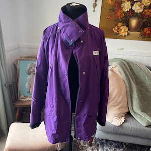 Puede incluir: Una chaqueta de color morado vibrante con cuello alto y cierre de botones a presión, cremallera frontal y dos bolsillos grandes. Un pequeño parche rectangular con el texto "LIFETIME DESIGN" es visible en la parte superior izquierda del pecho.