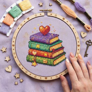 May include: Cross-stitch embroidery of a stack of books with a heart on top, inside a wooden hoop. The books are purple, green, orange, and green. Wooden stars, hearts, and a key are scattered around the hoop, along with colorful embroidery floss.
