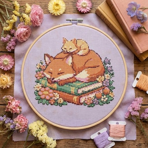 Puede incluir: Bordado de punto de cruz de dos zorros durmiendo sobre libros, rodeados de flores. Los zorros son de tonos naranja y crema, con los ojos cerrados. El bordado está en un aro de madera, con carretes de hilo y flores secas.