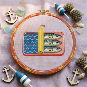 May include: A cross-stitch embroidery in a wooden hoop depicts a sardine can. The can is red and pink, with blue waves and three fish. The hoop is decorated with fish and red and yellow accents. Nautical theme.