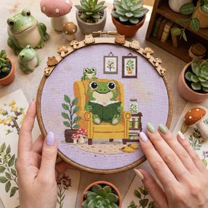 Op de afbeelding: Een borduurring met kruissteek met een kikker die een boek leest in een gele fauteuil. Het ontwerp omvat een kleine boekenkast, planten en ingelijste foto's. De ring is versierd met houten kikker- en paddenstoelversieringen.