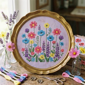 Op de afbeelding: Een geborduurd bloemenontwerp in kruissteek, omlijst door een goudkleurige, geschulpte lijst. Het ontwerp toont bloemen in roze, blauw, geel en paars. Borduurgaren en extra bloemen zijn rondom het frame geplaatst.