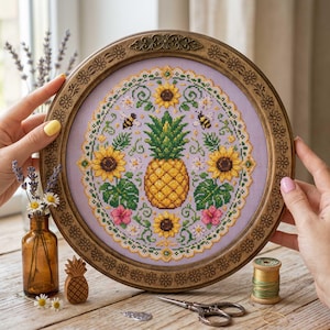 Peut inclure: Broderie au point de croix circulaire encadrée, représentant un ananas jaune, des tournesols, des abeilles et des feuilles tropicales sur fond lavande. Le cadre est en bois foncé avec des détails floraux. Décoration.