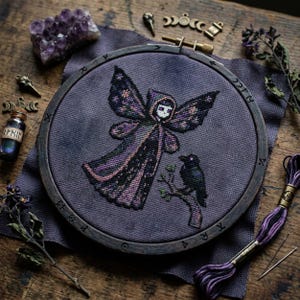 May include: A cross-stitch embroidery in a wooden hoop features a hooded figure with wings and a raven perched on a branch. The design uses shades of purple, black, and green on a gray background. Additional items include amethyst crystals and a small bottle.