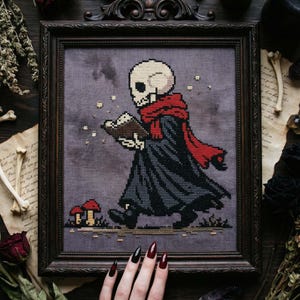 May include: Cross-stitch art in a dark wooden frame. The artwork depicts a skeleton figure in a black robe and red scarf, reading a book. Two red mushrooms are in the foreground. The background is a muted gray.