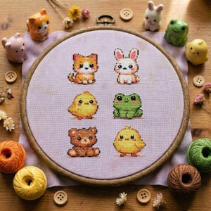 May include: A cross-stitch embroidery hoop featuring six cute animal designs: a cat, bunny, chick, frog, bear, and chick. The embroidery is set on a lavender fabric, surrounded by a wooden hoop. Decorative figurines and sewing supplies are arranged around the hoop.