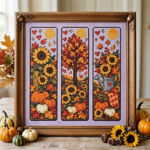 May include: A framed cross-stitch embroidery featuring three vertical panels depicting autumn scenes with sunflowers, orange and white pumpkins, a tree with fall foliage, and a blue watering can, all set against a light purple fabric background.