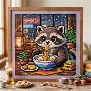 May include: A cross-stitch picture shows a raccoon eating ramen in a restaurant. The raccoon is brown and grey, with large eyes, eating noodles from a blue bowl. The scene includes a lantern, plants, and the Japanese word for ramen.