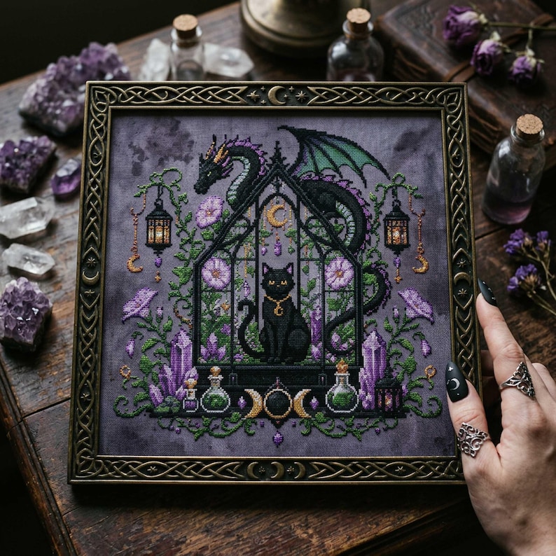 magical fantasy cross-stitch pattern featuring a black cat sitting inside a gothic greenhouse while a dark dragon with green wings curls protectively around the roof. The scene is decorated with lavender flowers, leafy vines, purple crystals, potion bottles, hanging lanterns, crescent moon charms, and celestial ornaments. The rich palette of black, green, lavender, gray, purple, and gold creates a mystical witchy garden mood, perfect for cat lovers, dragon fans, fantasy décor, and handmade walL