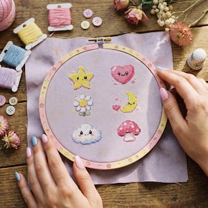 Pode incluir: Um bastidor de bordado com tecido lilás claro com desenhos de desenhos animados de uma estrela, um coração, uma flor, uma lua, uma nuvem e um cogumelo. O bastidor é segurado por mãos com unhas pintadas, rodeado de materiais de costura.