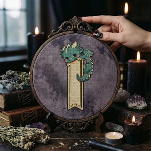 May include: A cross-stitch embroidery of a green dragon holding a banner, set in a dark wooden frame. The dragon is detailed with light green and pink accents. The background is a muted purple fabric. The frame is displayed on a wooden stand.