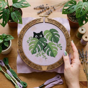以下が含まれることがあります： 木製のフレームに入ったクロスステッチ刺繍。黒猫が緑のモンステラの葉の間から覗いています。背景は薄紫色の布です。木製のフレームには葉の装飾模様があります。